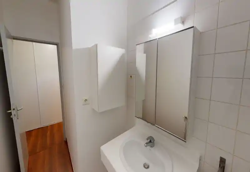 Salle de bains compacte avec lavabo mural, armoire à miroir et carreaux blancs; éclairage de la vasque lumineux et surfaces propres, adapté pour une annonce.