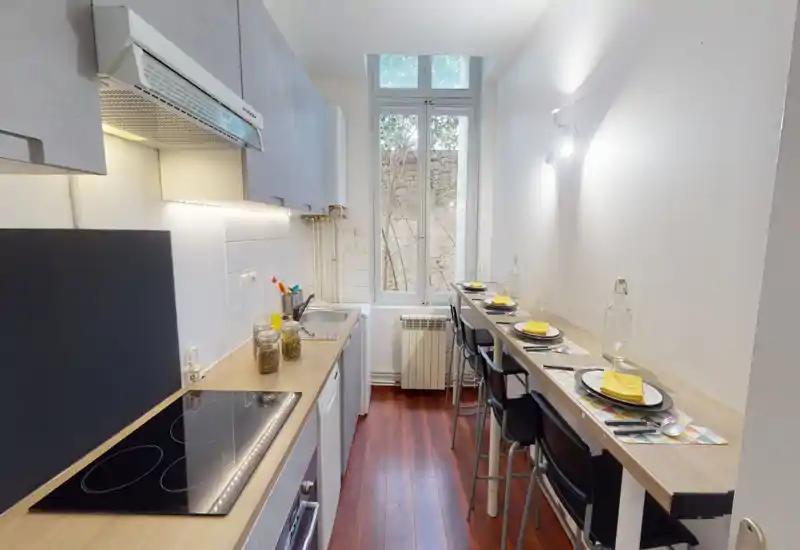 Cuisine compacte et moderne avec plaque à induction, évier et un bar petit-déjeuner pour quatre personnes. Meubles épurés, plans de travail en bois chaleureux et une fenêtre lumineuse créent un espace accueillant.