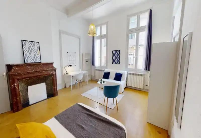 Camera da letto luminosa e ariosa con soffitti alti, letto matrimoniale visibile in primo piano, area salotto accogliente vicino alle finestre alte e dettagli decorativi eleganti come il camino d'epoca e la sospensione.