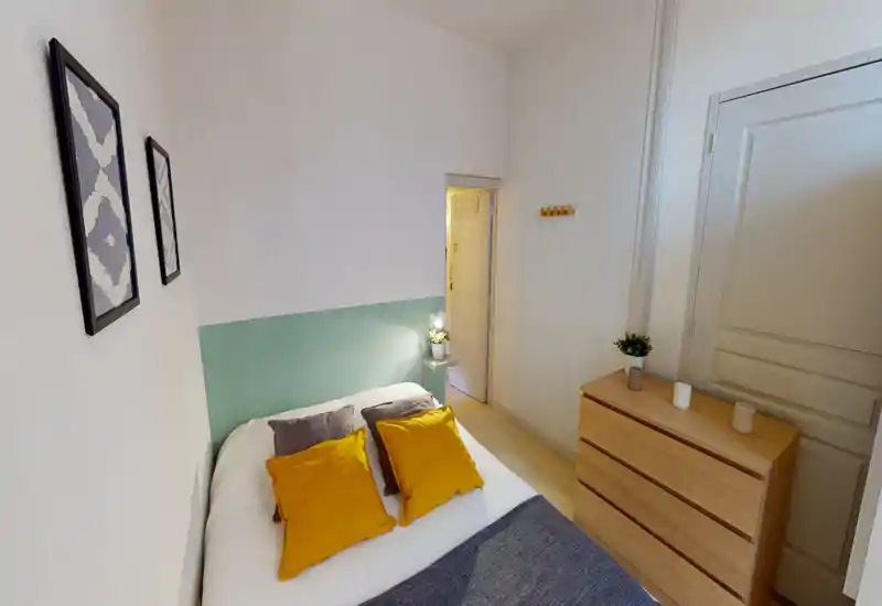 Camera da letto luminosa e ben arredata con letto matrimoniale in lenzuola bianche, cuscini color senape e un piccolo comò in legno accanto alla porta. Testiera pastello, quadretti a parete e lampada sul comodino donano un’atmosfera accogliente e moderna.