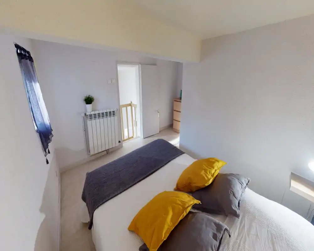 Bright, neatly staged bedroom with a double bed dressed in neutral linens, yellow accent pillows and a grey throw. The space feels compact and well-lit with a small window and simple furnishings including a dresser and radiator.