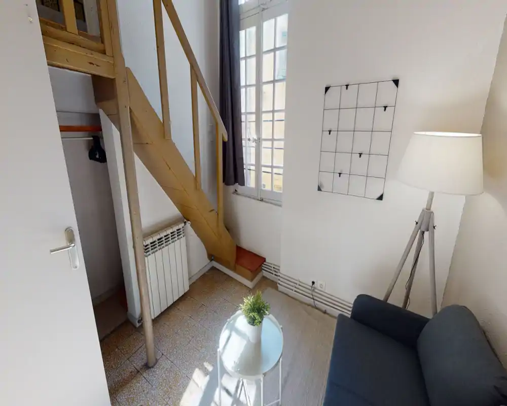 Compact living area with a small sofa, tripod floor lamp and a light round coffee table under natural window light — minimal, bright and functional.