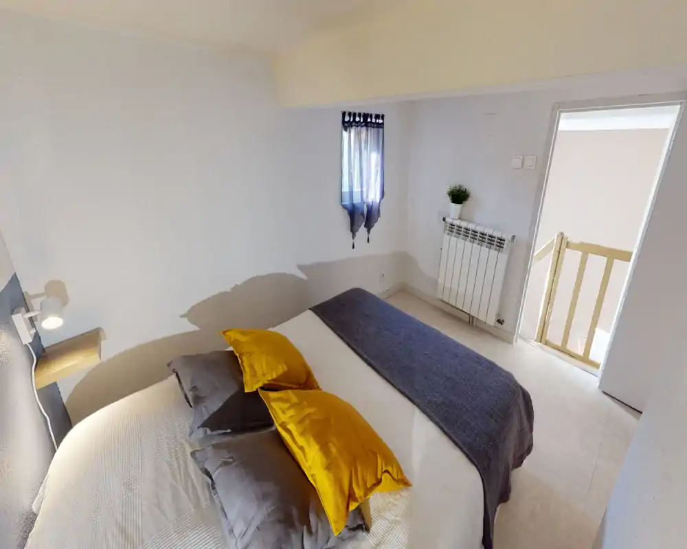 Bright, neatly styled bedroom with a fully visible double bed, warm accent pillows and a soft throw. The room features natural light from a small window and a simple bedside lamp for cozy evenings.