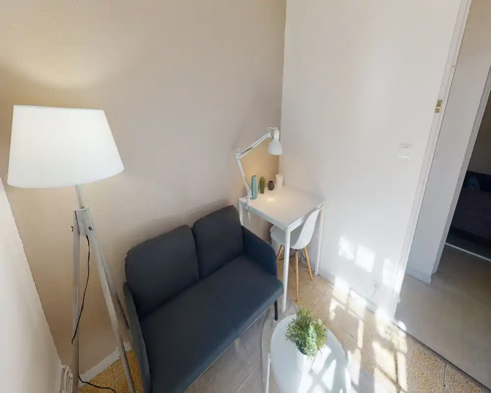 Bright, cozy living area with a compact dark sofa, tripod floor lamp, small white coffee table with plant and a corner desk — ideal for relaxing or working in a compact studio.