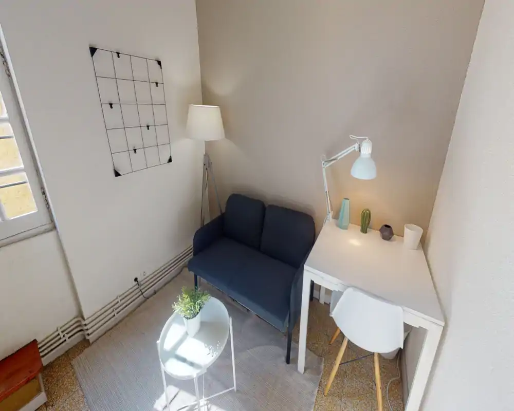 Compact, well-lit living area with a two-seater sofa, side table and a small desk; neutral tones and decorative lamp create a cozy, functional corner ideal for relaxing or working.