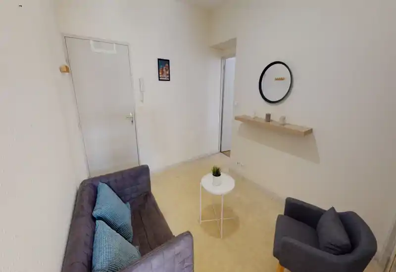 Zona giorno compatta con un piccolo divano, poltrona e tavolino tondo — stile moderno minimal con mensola decorativa e specchio. Pareti chiare e arredi semplici creano un ambiente ordinato e accogliente, ideale per rilassarsi.