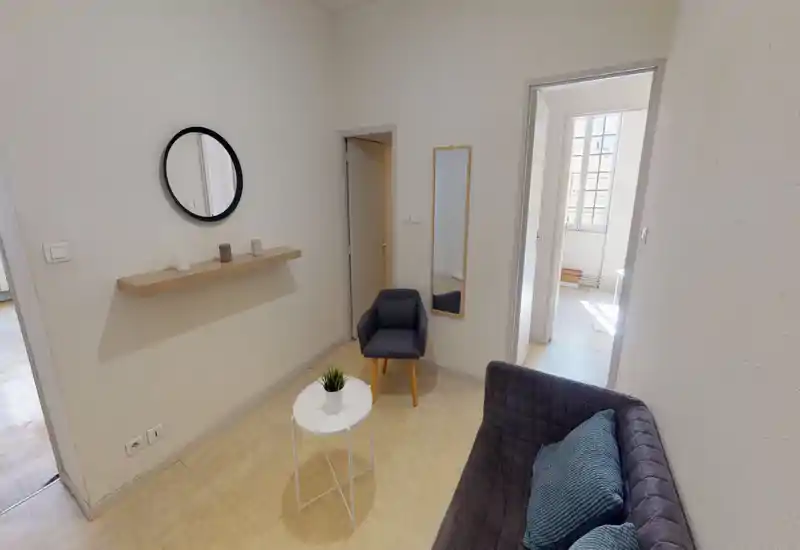 Piccolo soggiorno luminoso con divano trapuntato, poltrona e tavolino bianco. Arredamento minimal in stile scandinavo con mensola a parete, specchio rotondo e luce naturale dalla stanza adiacente.