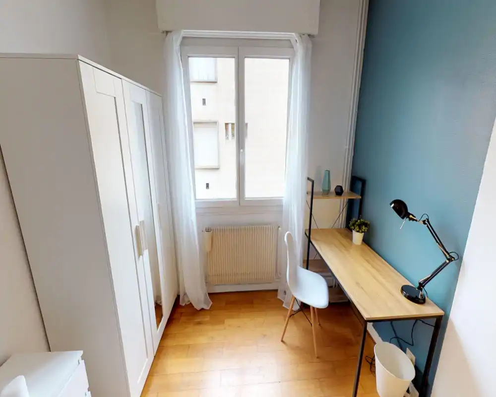 Small bedroom with a bright window, wooden floor and a neat study corner with desk, lamp and chair. White wardrobe and soft curtains create a tidy, airy atmosphere.