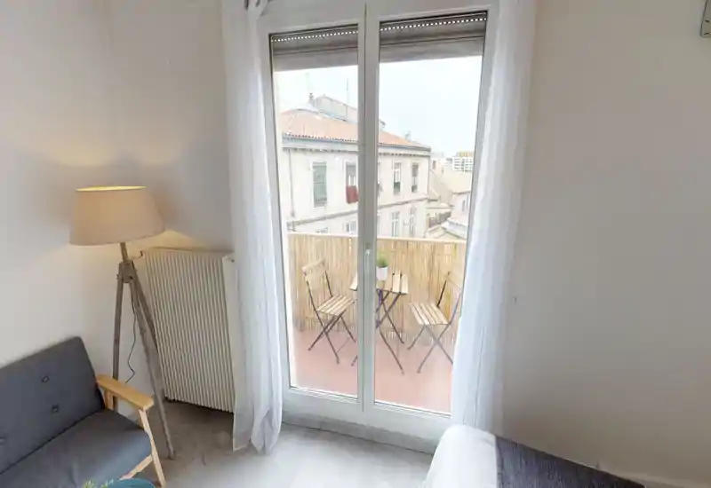 Bright bedroom corner with large glass doors opening to a small balcony. The shot shows part of the bed, a cozy floor lamp, a compact sofa and a bistro set outside, ideal for morning coffee with city views.