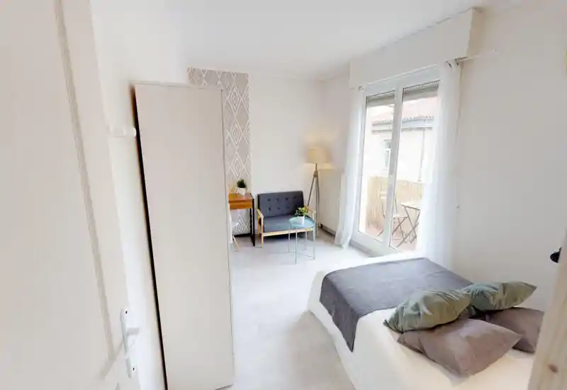 Sunlit, modern bedroom with a neatly made double bed, soft cushions and a small seating corner by the window that opens to a balcony. Bright neutral decor and simple furnishings create a calm, contemporary feel ideal for short-term rentals.