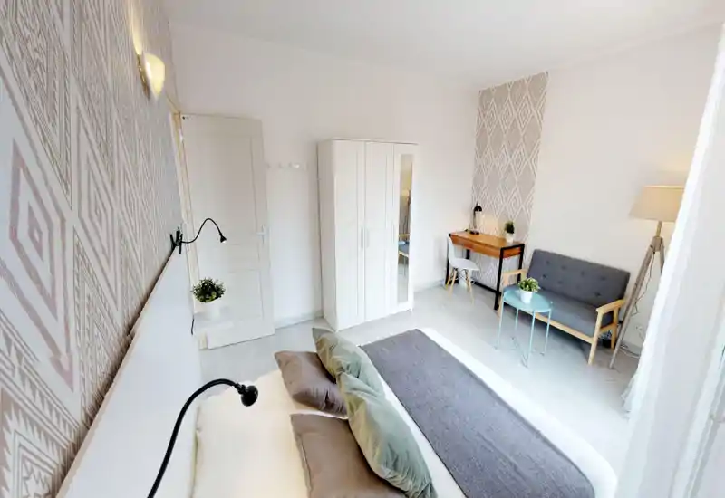 Bright, stylish bedroom with a comfortable double bed, decorative pillows and patterned wallpaper. A small seating area, desk and wardrobe make this room functional for work and relaxation.