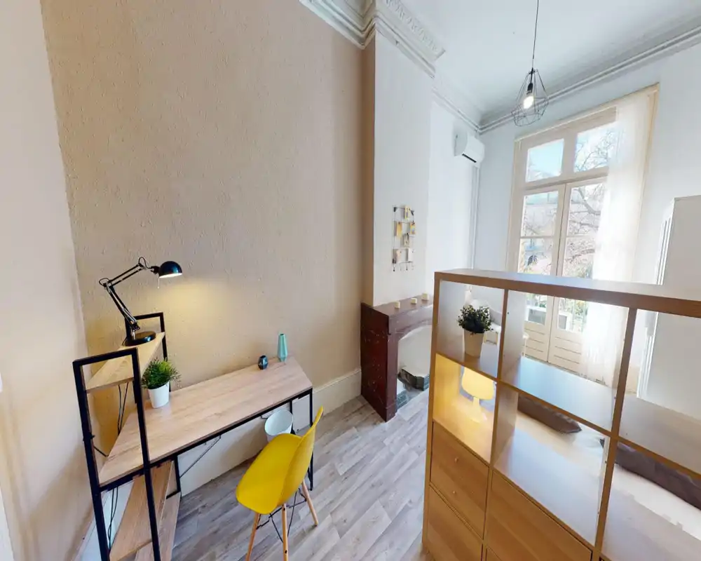 Coin chambre lumineux et tendance avec un bureau; le lit est partiellement visible derrière une étagère séparatrice. De grandes fenêtres apportent une belle lumière naturelle, l’espace est moderne et accueillant.