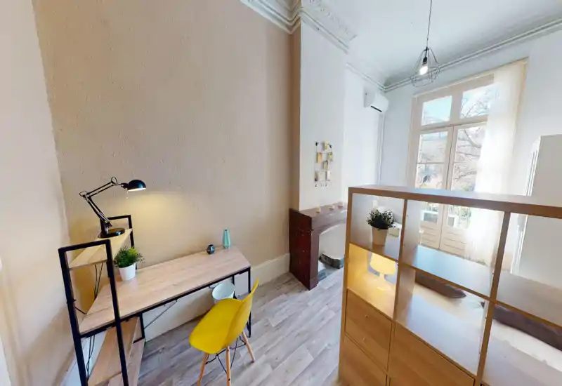 Coin chambre lumineux et tendance avec un bureau; le lit est partiellement visible derrière une étagère séparatrice. De grandes fenêtres apportent une belle lumière naturelle, l’espace est moderne et accueillant.
