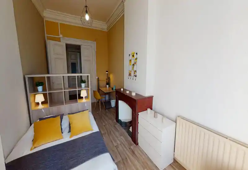 Chambre lumineuse et chaleureuse avec un lit double, des accents jaunes et une étagère tête de lit qui sépare l’espace nuit d’un petit bureau. Sol en bois, cheminée décorative et décoration soignée rendent la pièce accueillante.