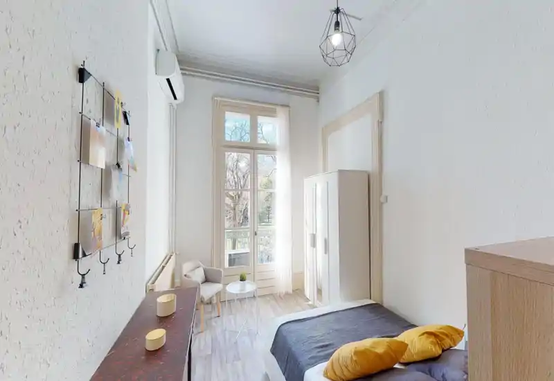 Chambre lumineuse et aérée avec grande fenêtre et vue sur balcon, lit double en linge neutre et coussins jaunes, armoire et petit coin salon — parfaite pour un séjour citadin ensoleillé.