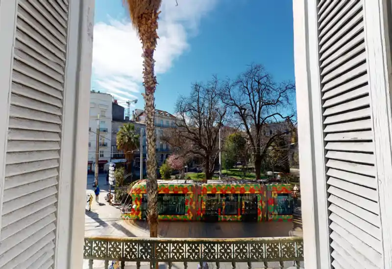 Vue ensoleillée depuis un balcon sur un tramway coloré et un petit parc urbain, encadrée par des volets classiques — parfaite pour mettre en valeur l'emplacement et l'animation de la rue.