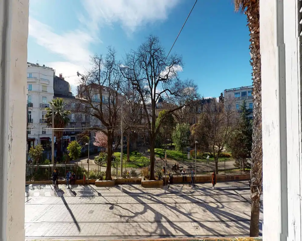 Vue ensoleillée sur un parc public depuis une fenêtre ou un balcon, avec de grands arbres, des allées et des bâtiments alentour sous un ciel bleu.