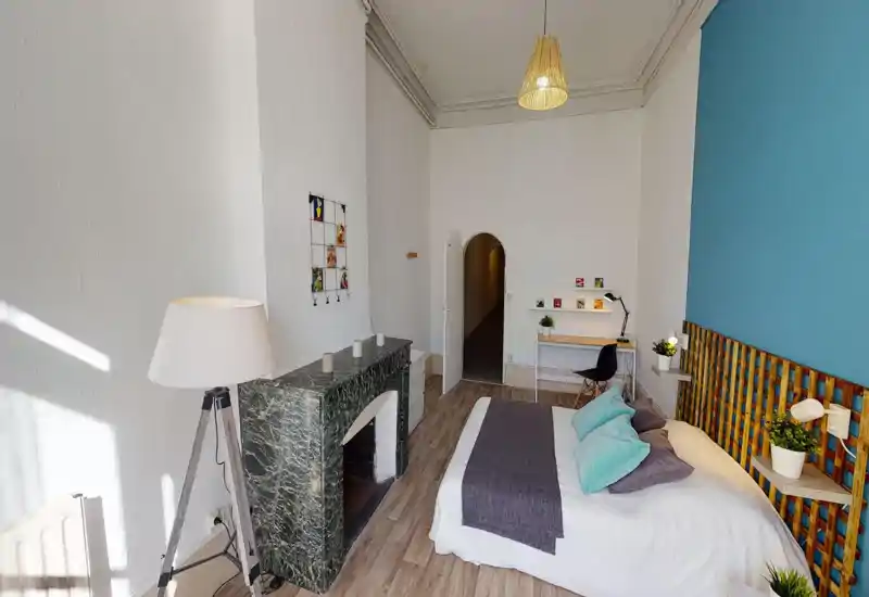 Chambre lumineuse et bien décorée avec un lit double confortable, une tête de lit texturée, des plantes de chevet et un petit coin bureau — idéale pour le repos et le travail.