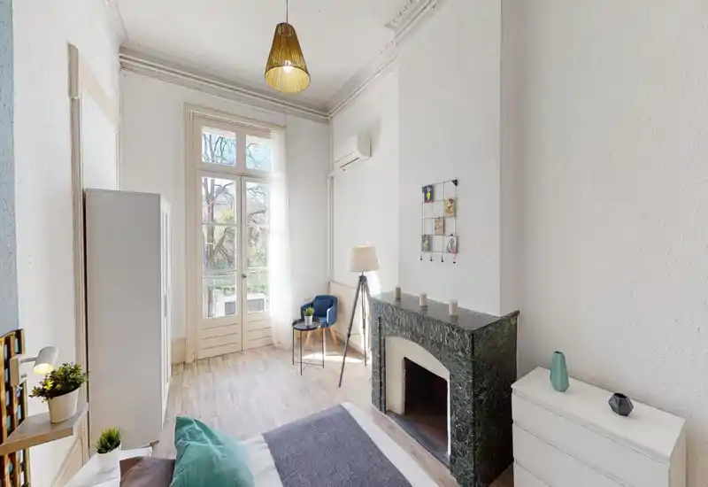 Chambre lumineuse et aérée avec une grande fenêtre et une décoration minimale soignée. Le lit est bien visible au premier plan, complété par une cheminée décorative, un fauteuil et une belle lumière naturelle.