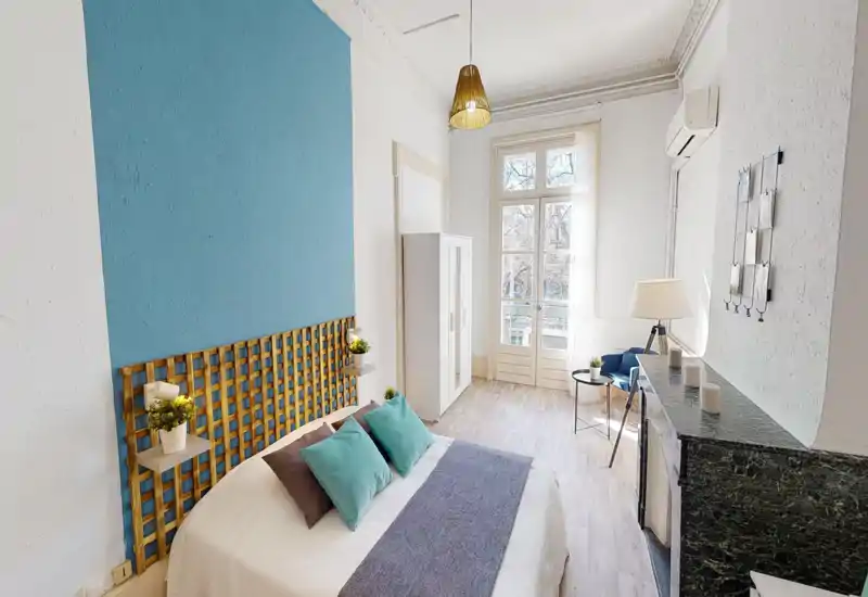 Chambre lumineuse et stylée avec un lit double, coussins décoratifs et une grande porte-fenêtre apportant une belle lumière naturelle. L’espace comprend une armoire, un coin détente et des accents en bleu et bois.