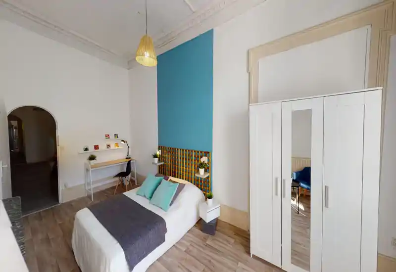 Chambre lumineuse et bien aménagée avec un lit double contre un mur bleu, textiles doux et une armoire blanche. Un petit bureau et des plantes décoratives apportent fonctionnalité et convivialité.