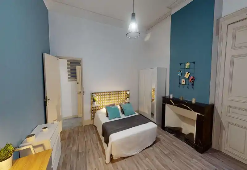 Chambre lumineuse et élégante avec lit double central, coussins turquoise, tête de lit éclairée et armoire. Sol effet bois et cheminée en marbre apportent du cachet.