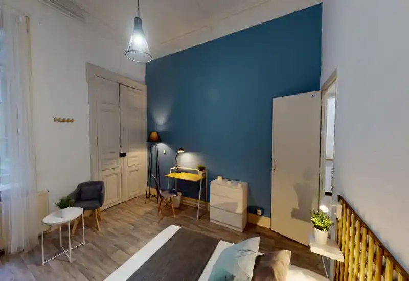 Chambre lumineuse et élégante avec un lit double visible au premier plan, mur d'accent bleu, lampes de chevet et petit bureau — confortable et bien éclairée.