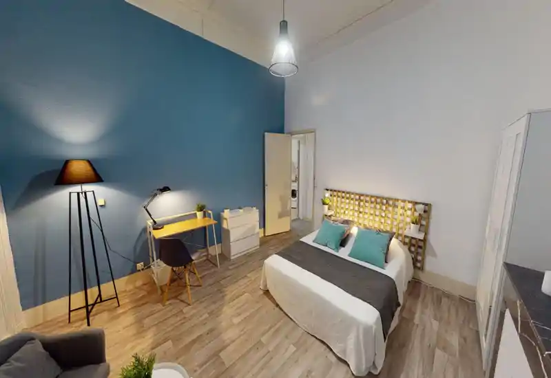 Chambre lumineuse et élégante avec lit double, tête de lit décorative et coussins bleu-vert coordonnés. La pièce dispose d'un bureau, d'une armoire et d'un éclairage d'ambiance chaleureux.