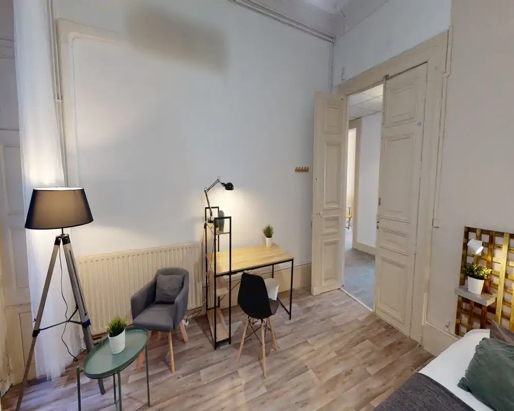 Bright, stylish bedroom corner with a small desk, armchair and floor lamp; a portion of the bed is visible at the right edge, making this a comfortable sleeping area with a functional workspace.