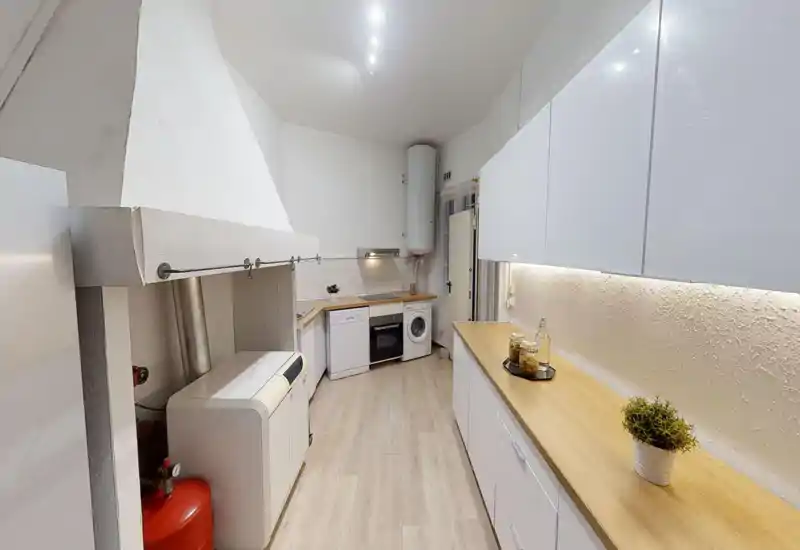 Cuisine en enfilade moderne avec meubles blancs, plans de travail en bois et appareils intégrés ; lumineuse, propre et bien éclairée, agrémentée de bocaux et plantes décoratives.
