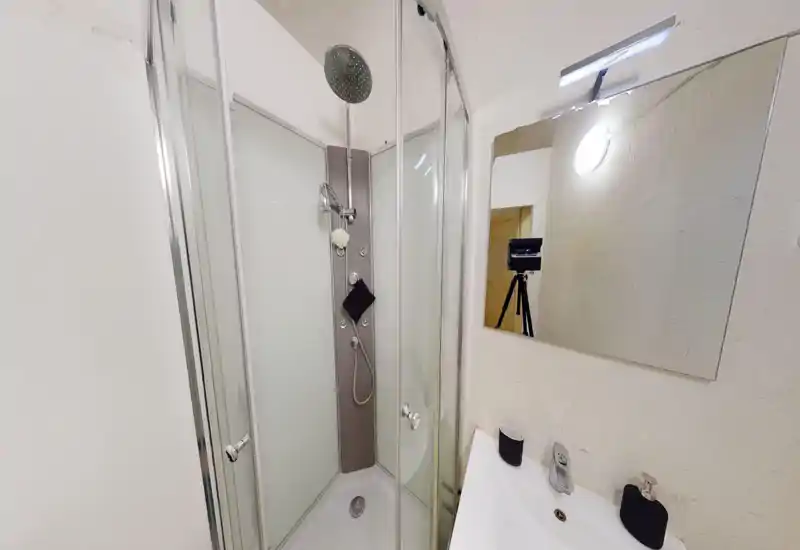Salle de bains compacte avec cabine de douche d'angle en verre, colonne de douche moderne et petit lavabo avec miroir — espace lumineux et fonctionnel adapté à un court séjour.