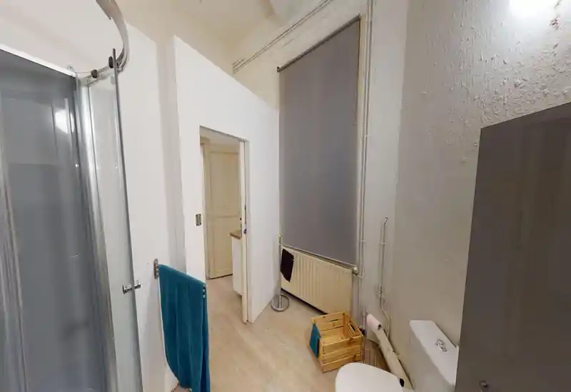 Salle de bains avec cabine de douche d'angle, WC, radiateur et sanitaires simples ; sol effet bois clair et murs neutres pour un espace fonctionnel et compact.