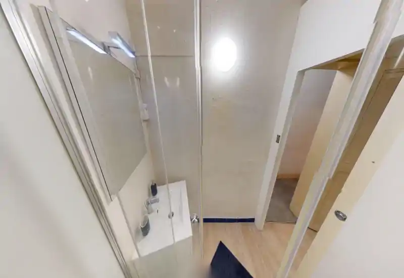 Salle de bain compacte avec petit lavabo, miroir et cabine de douche. Murs clairs et sol imitation bois pour un espace propre et fonctionnel.