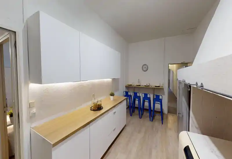 Cuisine moderne et lumineuse avec placards blancs, plan de travail en bois et petit bar avec tabourets bleus ; propre, bien éclairée et idéale pour des locations de courte durée.