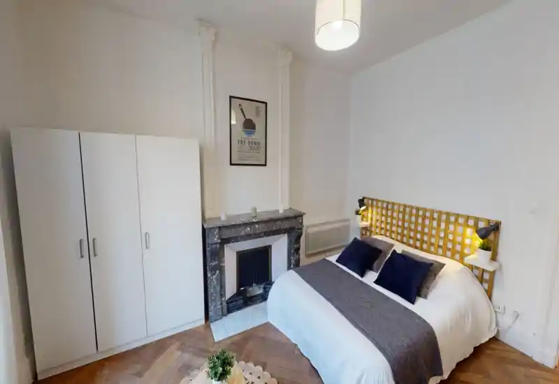 Bright, stylish bedroom with a double bed dressed in white linen and navy cushions, a decorative wooden headboard, bedside lamps and a wardrobe. The room features a marble fireplace and parquet flooring, creating a cozy, well-styled sleeping space ideal for rental listings.