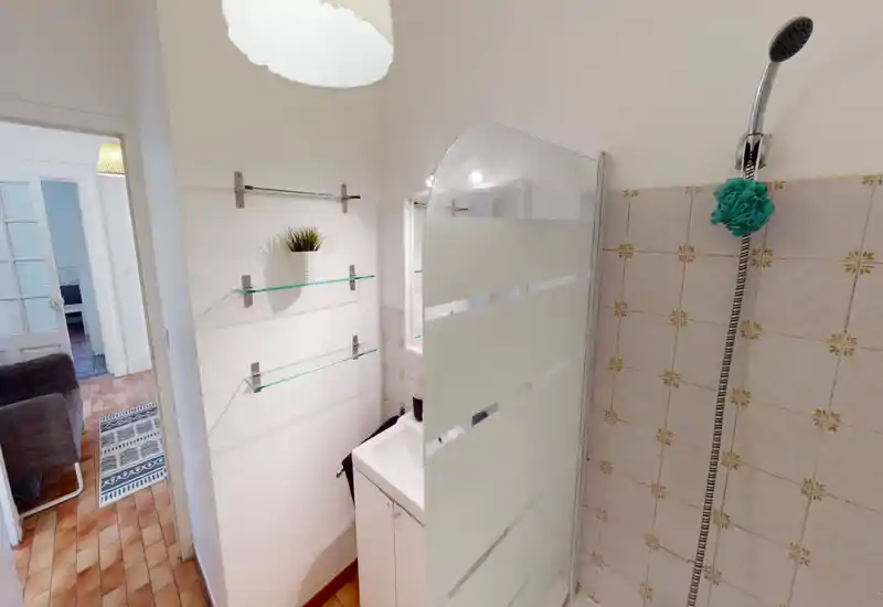 Salle de bain compacte avec douche murale, paroi de douche en verre dépoli et petit meuble vasque. Carrelage neutre et étagères simples pour un espace fonctionnel et lumineux.