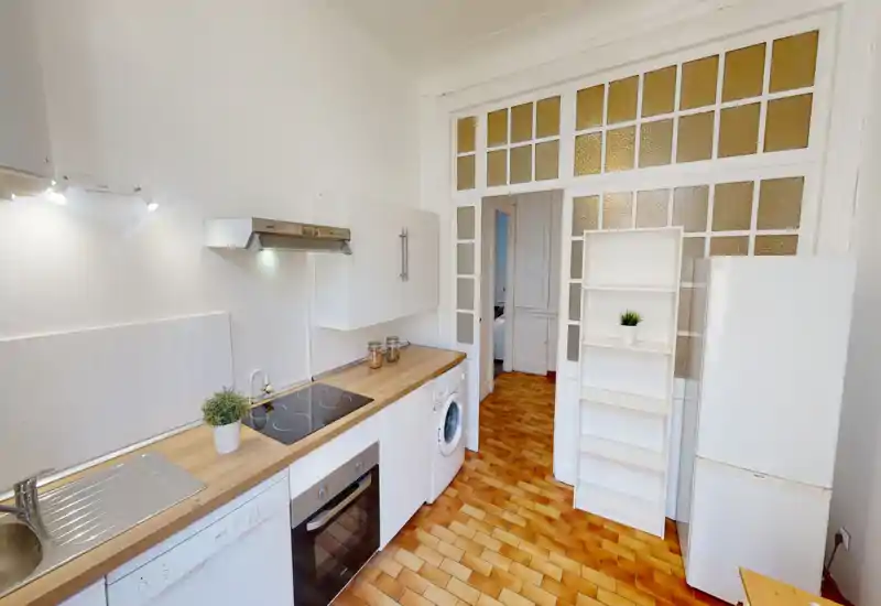 Cuisine compacte et lumineuse avec plan de travail en bois, plaque électrique, four encastré, lave-linge et grand réfrigérateur ; meubles blancs et sol en terre cuite apportent une atmosphère propre et chaleureuse.