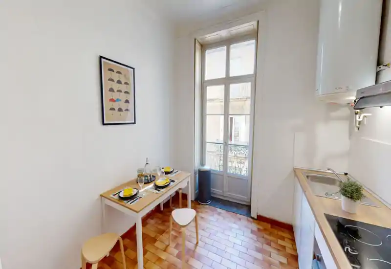 Cuisine compacte et lumineuse avec plan de travail en bois, évier et plaque à induction. Une petite table dressée pour deux se trouve près d'une grande fenêtre-fenêtre laissant entrer la lumière naturelle.
