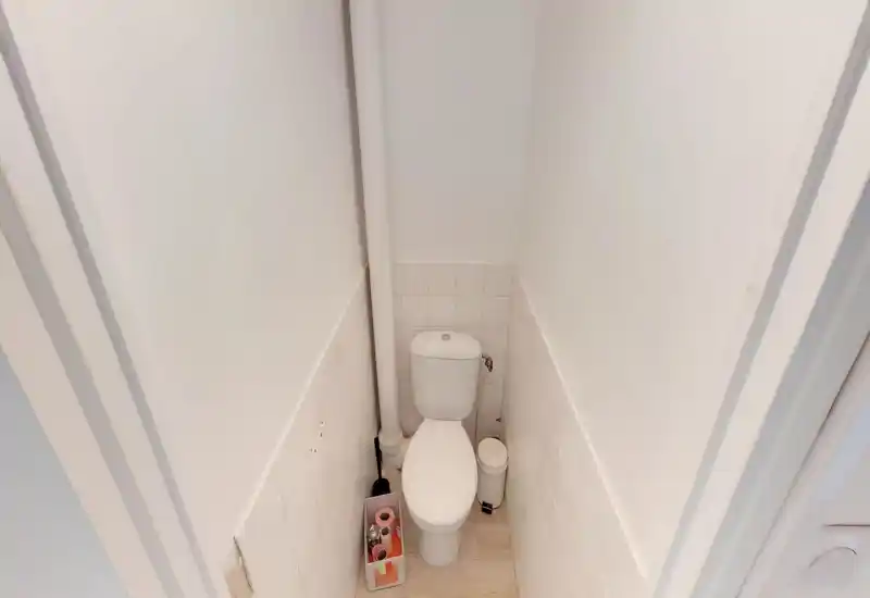 WC compact avec cuvette blanche adossée à des demi-murs carrelés, une colonne d'évacuation verticale et petits accessoires (poubelle, brosse, rouleaux). Lumineux, simple et propre.