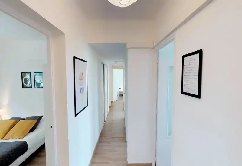 Couloir lumineux et étroit avec sol en bois clair et cadres muraux menant aux pièces adjacentes ; entrée moderne et épurée donnant sur une chambre.