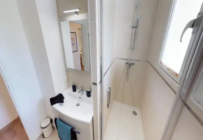 Salle de bain compacte et moderne avec douche, mitigeur mural, lavabo et miroir, accessoires rangés. Carrelage neutre et lumière naturelle provenant d'une fenêtre dépoli pour une impression de propreté.