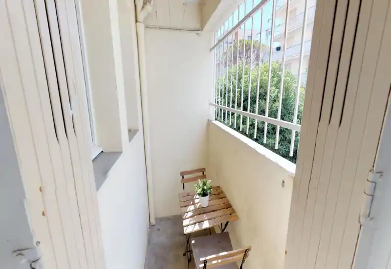 Petit balcon privé avec table bistro en bois et deux chaises, barrières métalliques et vue végétale ; lumineux et ensoleillé, parfait pour un café du matin.