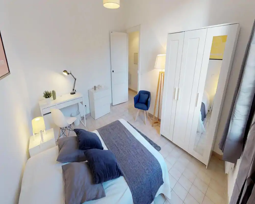 Bright, well-staged bedroom with a double bed dressed in neutral linens, soft pillows and a textured throw; a white wardrobe with mirror, desk and a cozy armchair complete the space.