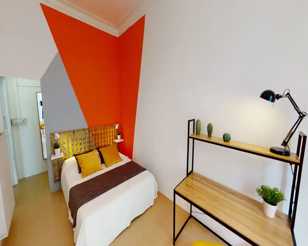 Chambre moderne et lumineuse avec un lit double devant un mur accent orange géométrique, coussins assortis et un bureau en bois avec lampe et plantes.