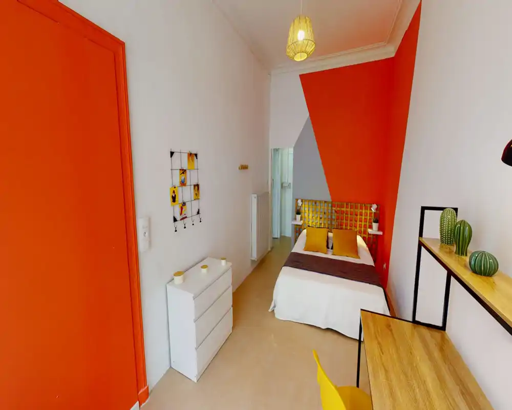 Chambre lumineuse et soignée avec un lit double central, mur d'accent orange et coussins assortis ; bureau et étagères offrent un espace de travail pratique.