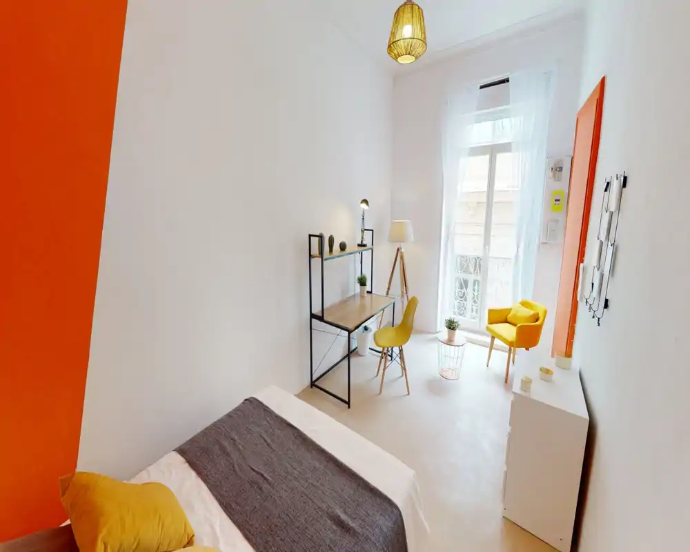 Chambre moderne et lumineuse avec un lit au premier plan, un bureau en bois minimaliste et un coin détente près d'une grande fenêtre laissant entrer beaucoup de lumière naturelle.