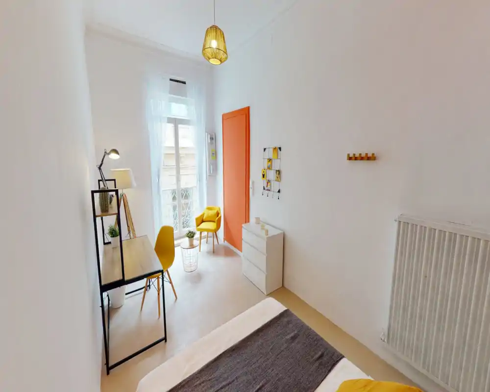 Chambre lumineuse et moderne avec lit visible au premier plan, un petit bureau et un fauteuil jaune chaleureux près d'une grande fenêtre laissant entrer beaucoup de lumière naturelle.
