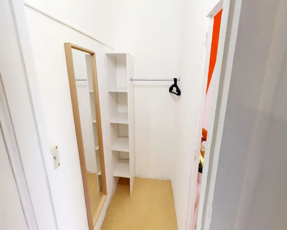 Petit dressing/entrée avec étagères intégrées, tringle pour vêtements et miroir en pied ; murs blancs et sol carrelé.