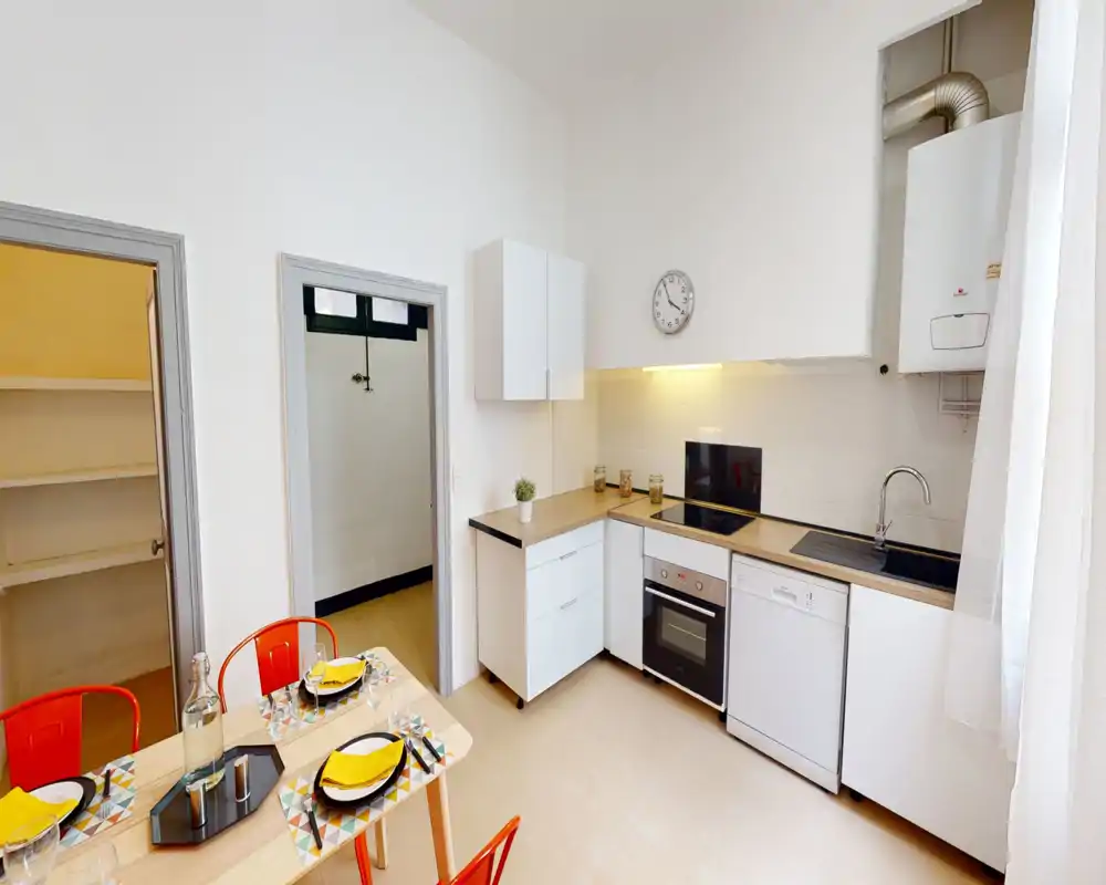 Cuisine moderne et lumineuse avec four encastré, lave-vaisselle et une petite table dressée. Placards blancs, plans de travail en bois et fenêtre avec rideau pour un espace net et accueillant.