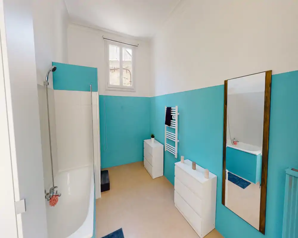Salle de bain lumineuse et propre avec baignoire et douchette, peinture turquoise sur la moitié des murs, commodes blanches et grand miroir à cadre bois.